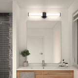 A modern bathroom with a large mirror, a wooden vanity with dual sinks, a potted plant, soap dispenser, and toiletries. Above the mirror is a sleek, horizontal light fixture. A tiled shower is visible to the left.