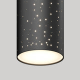 A close-up of a cylindrical black pendant light with small circular perforations, allowing light to shine through and create a star-like pattern. The main light source glows softly from the open bottom.