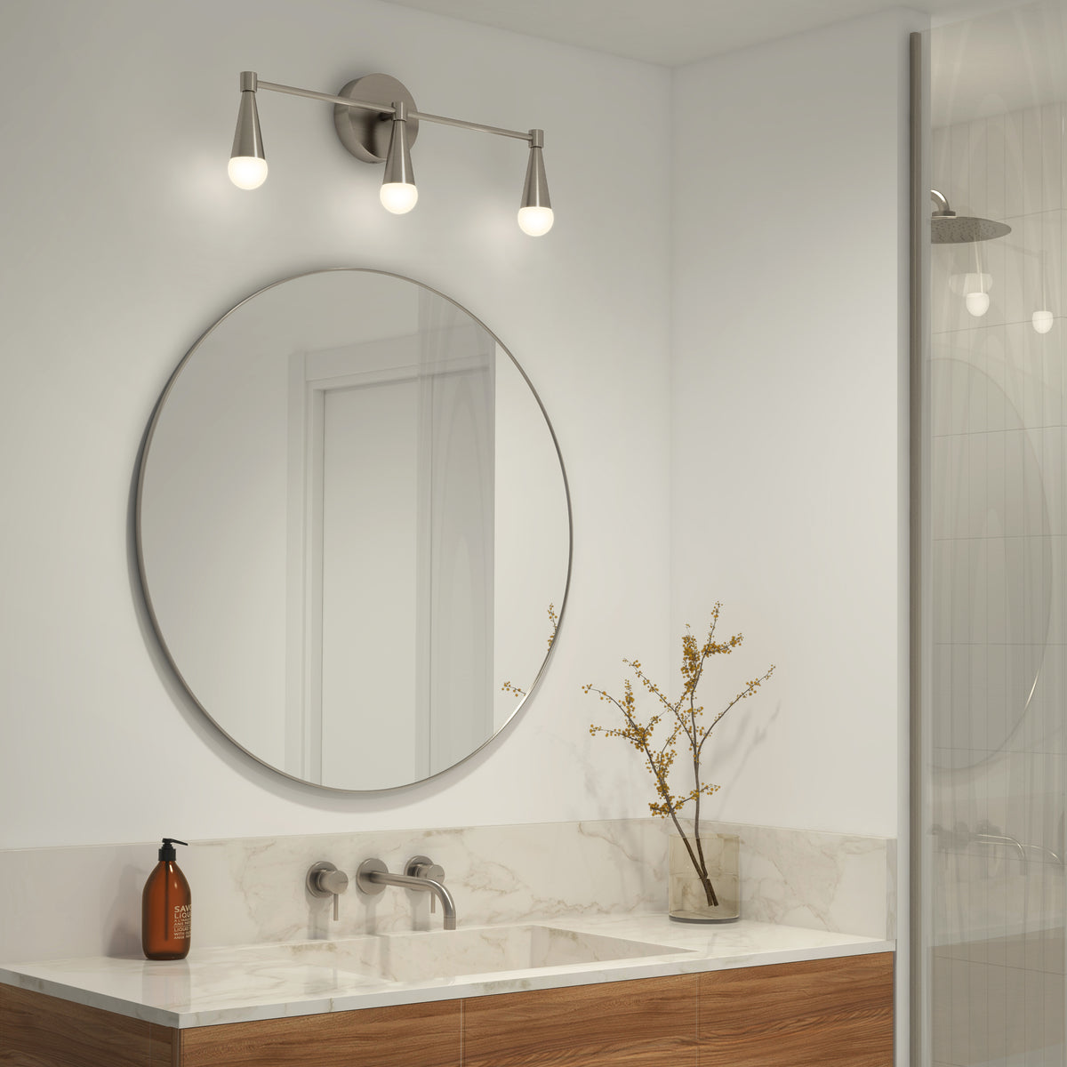 A modern bathroom with a round mirror above a marble countertop, wall-mounted faucet, a bottle of soap, and a vase with branches. Three light bulbs are mounted above the mirror, and a glass shower is visible on the right.