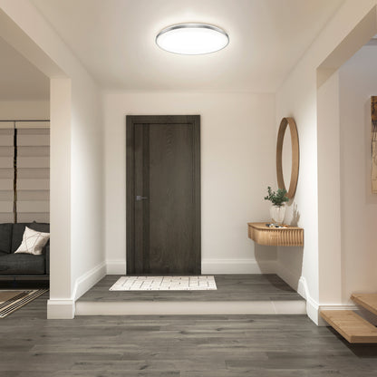 A modern entryway features a dark wooden door, grey wood flooring, a floating console with a mirror and plant, and the Artika Europa 21 in. LED modern flush mount light with remote overhead. A living room with a sofa is visible to the left.