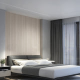 A modern bedroom with a gray upholstered bed, white bedding, and a gray throw. Behind the bed is a textured beige accent wall. Dark curtains and sheer drapes cover a large window; a potted plant sits on the bedside table.