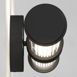 A modern black cylindrical wall sconce with a clear ribbed glass cover is mounted on a light-colored wall, emitting a bright light. The view is from above, highlighting its sleek and minimal design.