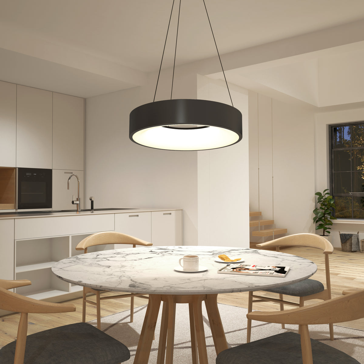 A modern kitchen and dining area with a round marble table, wooden chairs, a cup and magazine on the table, a large circular ceiling light above, white cabinetry, and stairs in the background.