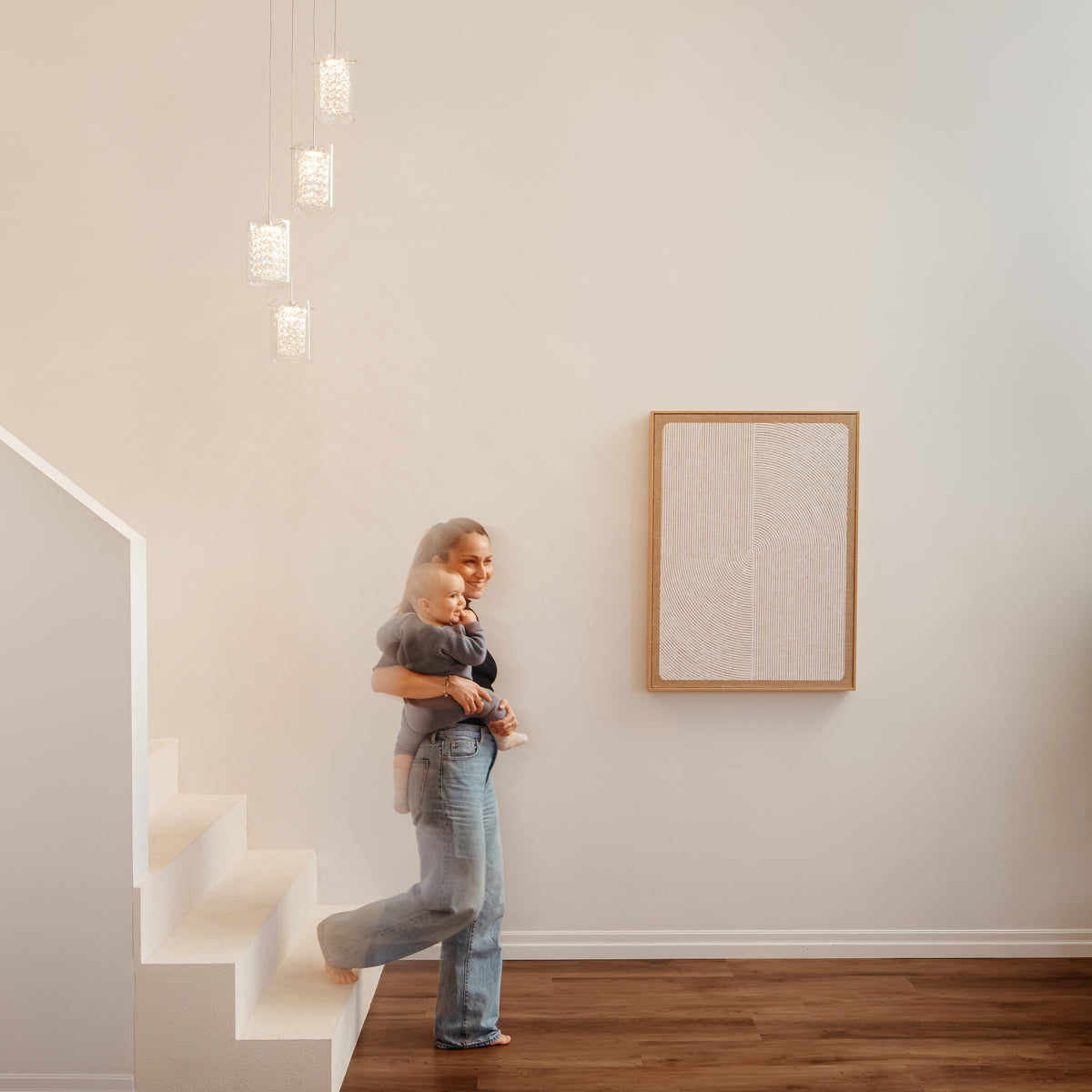 A woman wearing jeans stands at the base of a white staircase holding a young child. Modern pendant lights hang above, and abstract art is mounted on the white wall beside them. The floor is wooden.