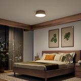A cozy modern bedroom with a wooden bed, earth-toned bedding, two botanical prints on the wall, a round ceiling light, and a night view of a city skyline visible through large windows with sheer curtains.