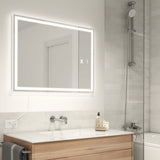 Modern bathroom with a large, illuminated rectangular mirror above a white sink and wooden vanity. A glass shower with a chrome showerhead is on the right. The walls are tiled in light gray.