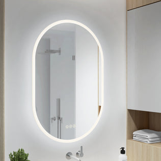 Artika’s Clara LED Mirror Oval 30x20″ with Defogger reflects a shower; shelves nearby hold towels and a soap dispenser.