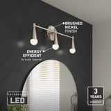 A modern three-light vanity fixture with brushed nickel finish and teardrop-shaped LED bulbs is mounted above a round mirror on a dark chevron-tiled wall; text highlights energy efficiency and 3-year warranty.