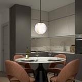 A modern kitchen with gray cabinets and a marble backsplash features a round table set for two, with wine glasses, a bottle of wine, and pink cushioned chairs under a spherical pendant light.
