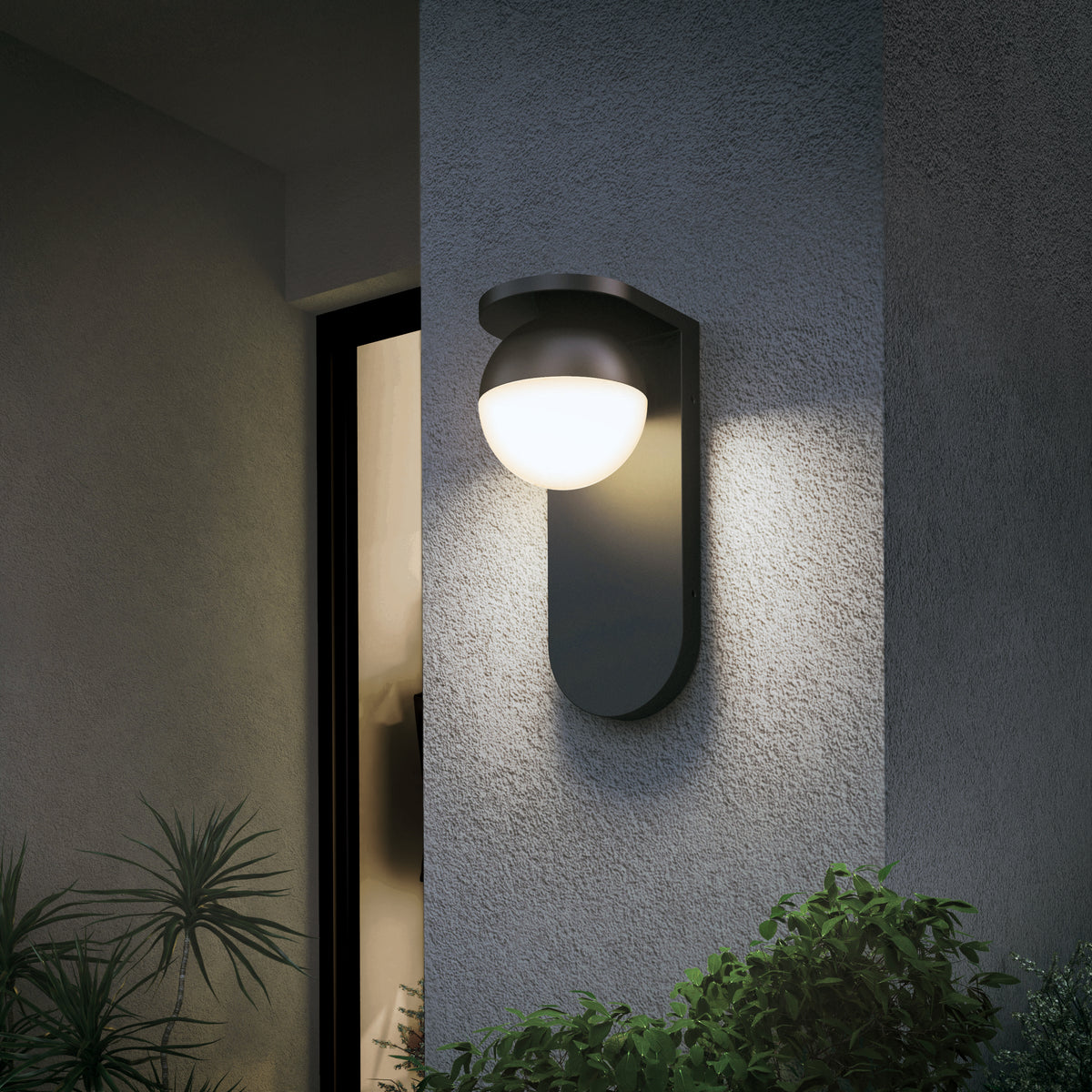A modern outdoor wall light with a round, frosted glass shade is mounted on a textured gray wall near a window, illuminating nearby green plants in the evening.