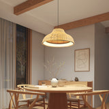 A cozy dining area with a round wooden table, four matching chairs, a woven pendant light, and a vase with branches. Soft light filters through sheer curtains, and minimalist art hangs on the wall.