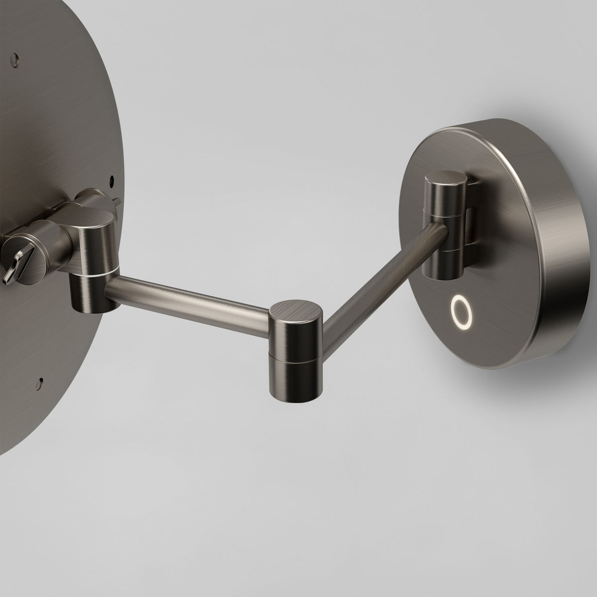 A close-up of a modern, metallic wall-mounted adjustable mirror arm with round joints and a circular wall plate, set against a plain light gray background.