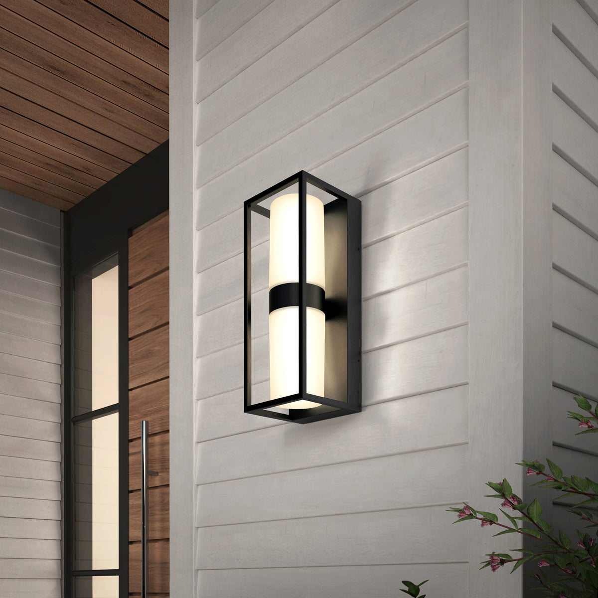 Modern black-framed outdoor wall sconce with cylindrical frosted glass, mounted on a light gray horizontal-paneled exterior wall near a wooden door; some greenery is visible in the corner.