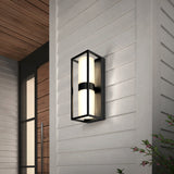 Modern black-framed outdoor wall sconce with cylindrical frosted glass, mounted on a light gray horizontal-paneled exterior wall near a wooden door; some greenery is visible in the corner.