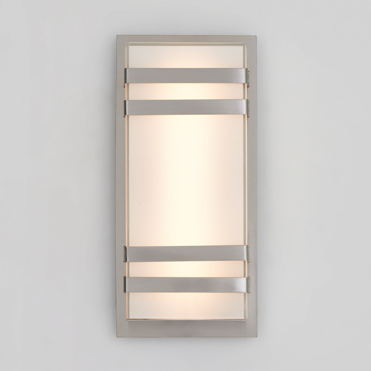 The Artika Glowbox 3 indoor-outdoor LED wall light features a frosted glass center with a metallic frame and three horizontal metal bands, casting soft, diffused light onto a light gray wall.