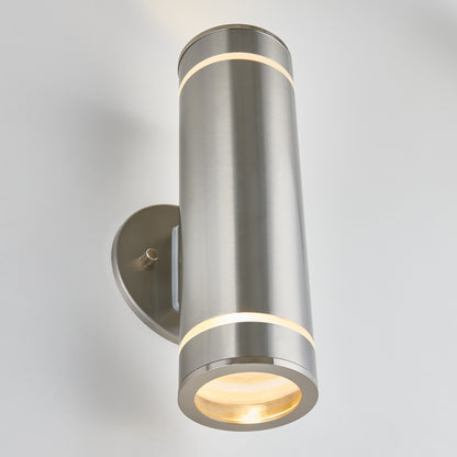 The Artika C7 indoor-outdoor wall light features a modern cylindrical brushed metal design, with illumination from both the top and bottom, mounted on a sleek oval wall plate against a white background.
