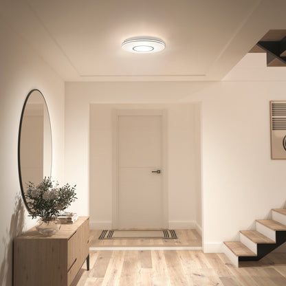 A modern hallway features light wood flooring, a round mirror above a wooden console with a plant, a white door, and stairs. The space is illuminated by the Artika Horizon 13 in. LED flush mount light 3 CCT on the ceiling.