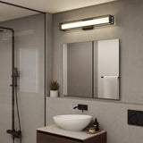 A modern bathroom with gray tiled walls, a rectangular mirror above a wooden vanity with a white vessel sink, black wall-mounted faucet, and a rectangular light fixture above the mirror. A small plant and toiletries are on the counter.