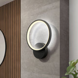 A modern circular wall sconce with a glowing LED ring mounted on a gray wall, next to a wooden slat accent and a plant in the lower right corner.