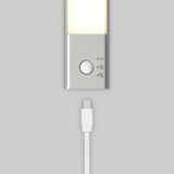 A close-up of a rectangular LED light with a motion sensor and three icons on its silver base, positioned above a white USB-C charging cable against a light gray background.