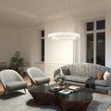 A modern living room with a gray velvet sofa, two matching armchairs, a black coffee table, a plush rug, and a large circular chandelier. The space features big windows, white walls, and decorative plants.