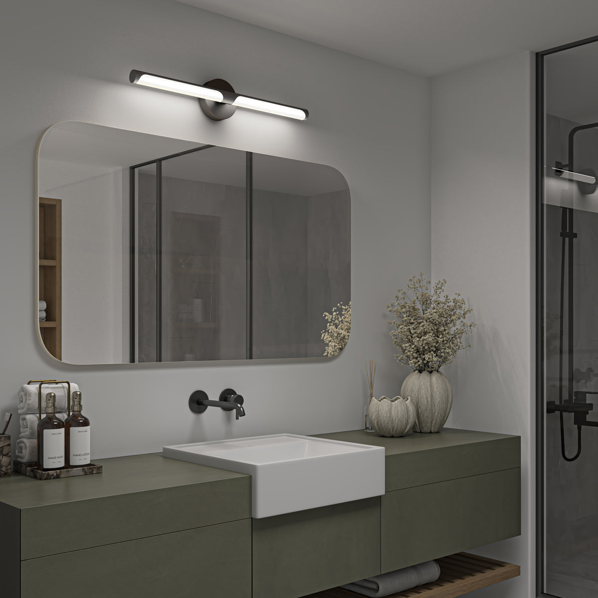 Modern bathroom with a large rectangular mirror, wall-mounted light above, minimalist sink, green vanity, decorative vases with dried flowers, toiletries, neatly folded towels, and a glass shower enclosure in the background.