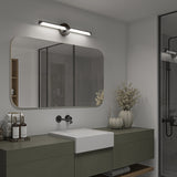 Modern bathroom with a large rectangular mirror, wall-mounted light above, minimalist sink, green vanity, decorative vases with dried flowers, toiletries, neatly folded towels, and a glass shower enclosure in the background.