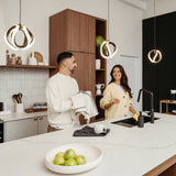 A man and woman smile at each other while washing and drying dishes together in a modern kitchen. There is a bowl of green apples on a white countertop and stylish pendant lights hang above.
