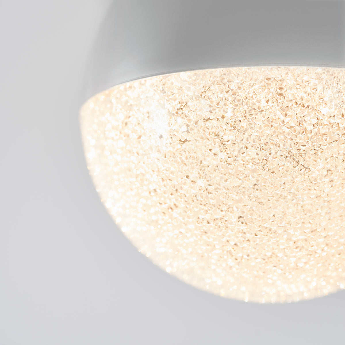 Close-up of a modern ceiling light fixture with a textured, sparkling glass or crystal dome that emits a warm, diffused glow. The background is plain and light-colored, highlighting the lamp’s design.