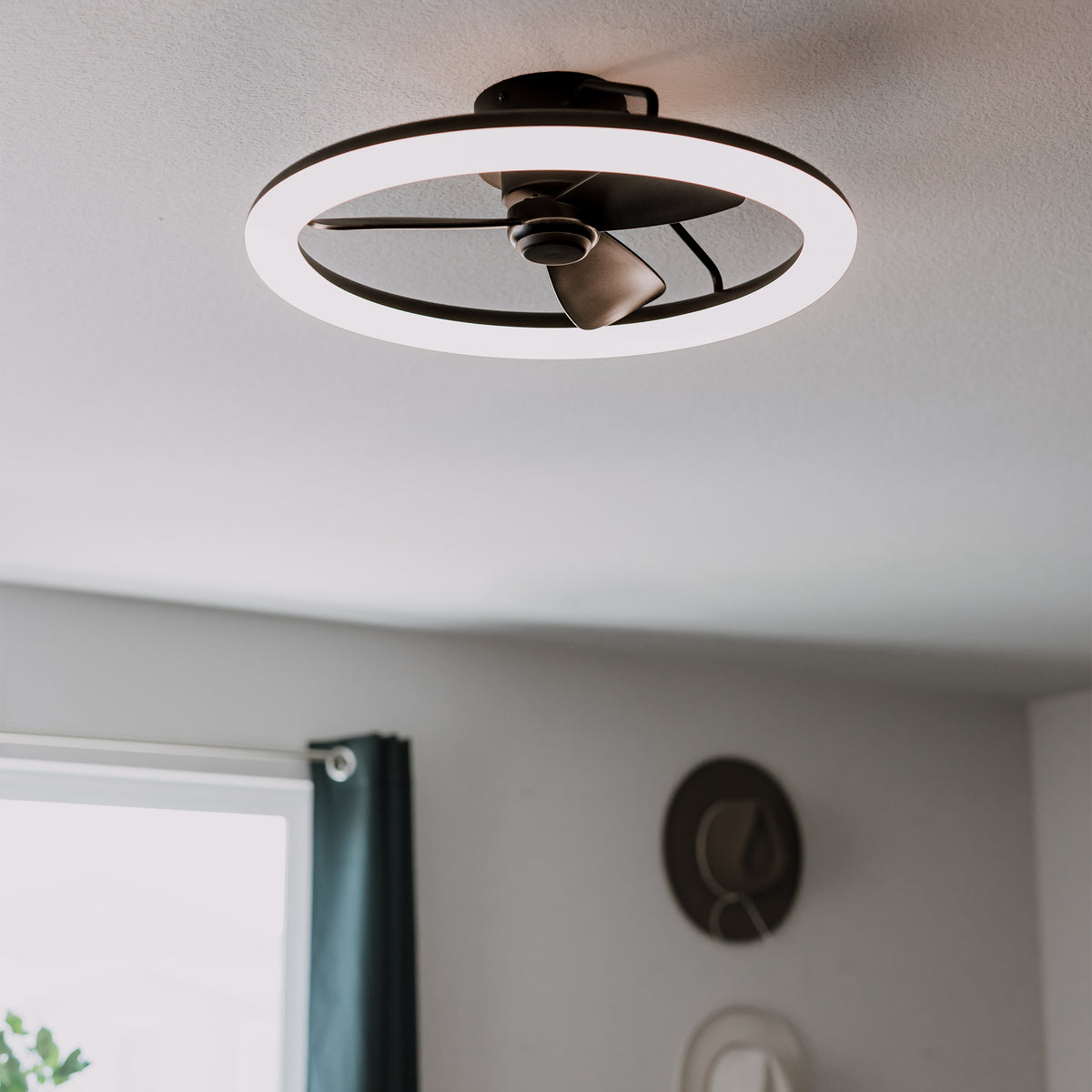 A modern ceiling fixture featuring a circular LED light with a small fan in the center, mounted on a white ceiling in a room with gray walls and a window with a dark curtain.
