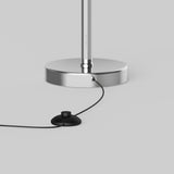 A close-up of the base of a chrome floor lamp with a black power cord and a round black foot switch on a light gray background.