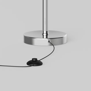 A close-up of the Artika Pivot modern LED floor lamp 3 CCT shows its chrome base with a connected black cord and round foot switch, displayed on a light gray surface.