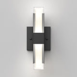 A modern wall sconce with a rectangular black base and two vertical light bars, emitting soft white light from the center. The design is minimalistic and geometric, mounted on a plain light gray wall.