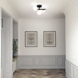 A minimalistic hallway features light gray paneling, two framed abstract art pieces on the wall, a potted plant on a windowsill, and a modern ceiling light with three round bulbs. The floor has a herringbone pattern.