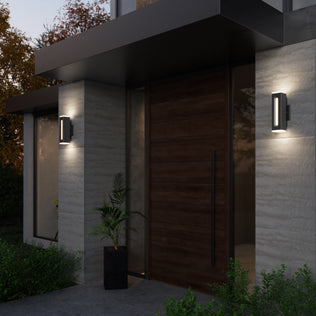 A modern house entrance at dusk features a large wooden door, gray walls, and Artika Linea Square LED indoor-outdoor wall lights on either side. A potted plant is near the door, with greenery surrounding the entryway.