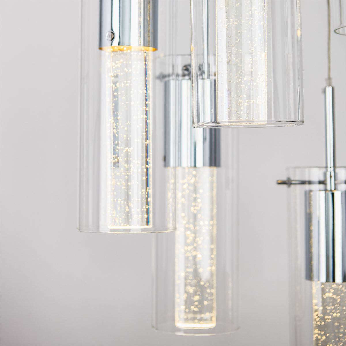 Close-up of modern cylindrical pendant lights with clear glass and chrome finishes, featuring tiny illuminated bubbles inside each tube, creating a sparkling effect against a neutral background.