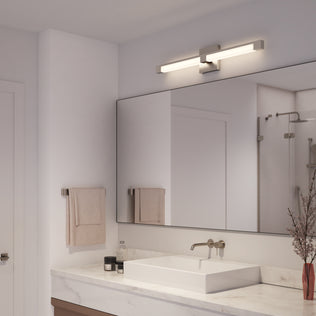 Modern bathroom featuring the Artika Tivoli modern LED bathroom vanity light (5 CCT) above a large rectangular mirror, white sink on marble countertop, beige towels, and a vase with dried branches. Shower visible in the reflection.