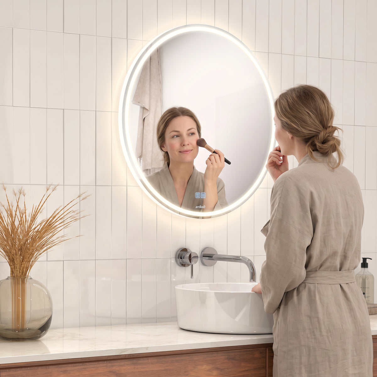 A woman applies makeup before the Artika Emeraude 24x24 in. Anti-Fog LED Bathroom Mirror, its glow lighting the vanity.