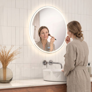 A woman applies makeup before the Artika Emeraude 24x24 in. Anti-Fog LED Bathroom Mirror, its glow lighting the vanity.