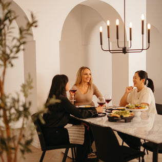 Three women enjoy red wine and conversation at a dining table set with food, while the Artika Manchester 6-light pendant casts a warm glow overhead in a stylish room with arched openings.