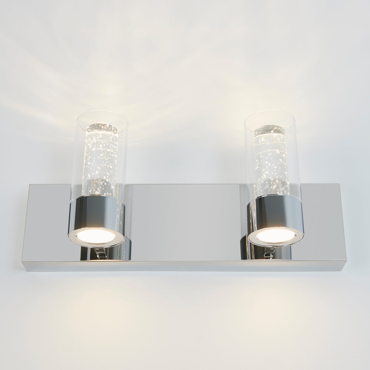 A modern wall sconce with a rectangular chrome base and two cylindrical glass shades, each containing small bubbles and lit from below, creating a sparkling effect on a white wall.