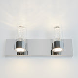A modern wall sconce with a rectangular chrome base and two cylindrical glass shades, each containing small bubbles and lit from below, creating a sparkling effect on a white wall.