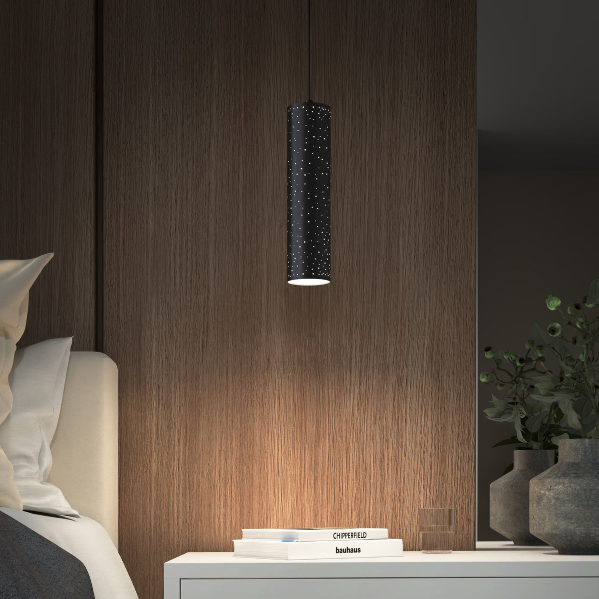 A modern bedroom features a cylindrical black pendant light with small holes, casting a soft glow over a white nightstand with stacked books, a glass of water, and a green leafy plant.