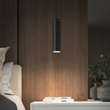 A modern bedroom features a cylindrical black pendant light with small holes, casting a soft glow over a white nightstand with stacked books, a glass of water, and a green leafy plant.