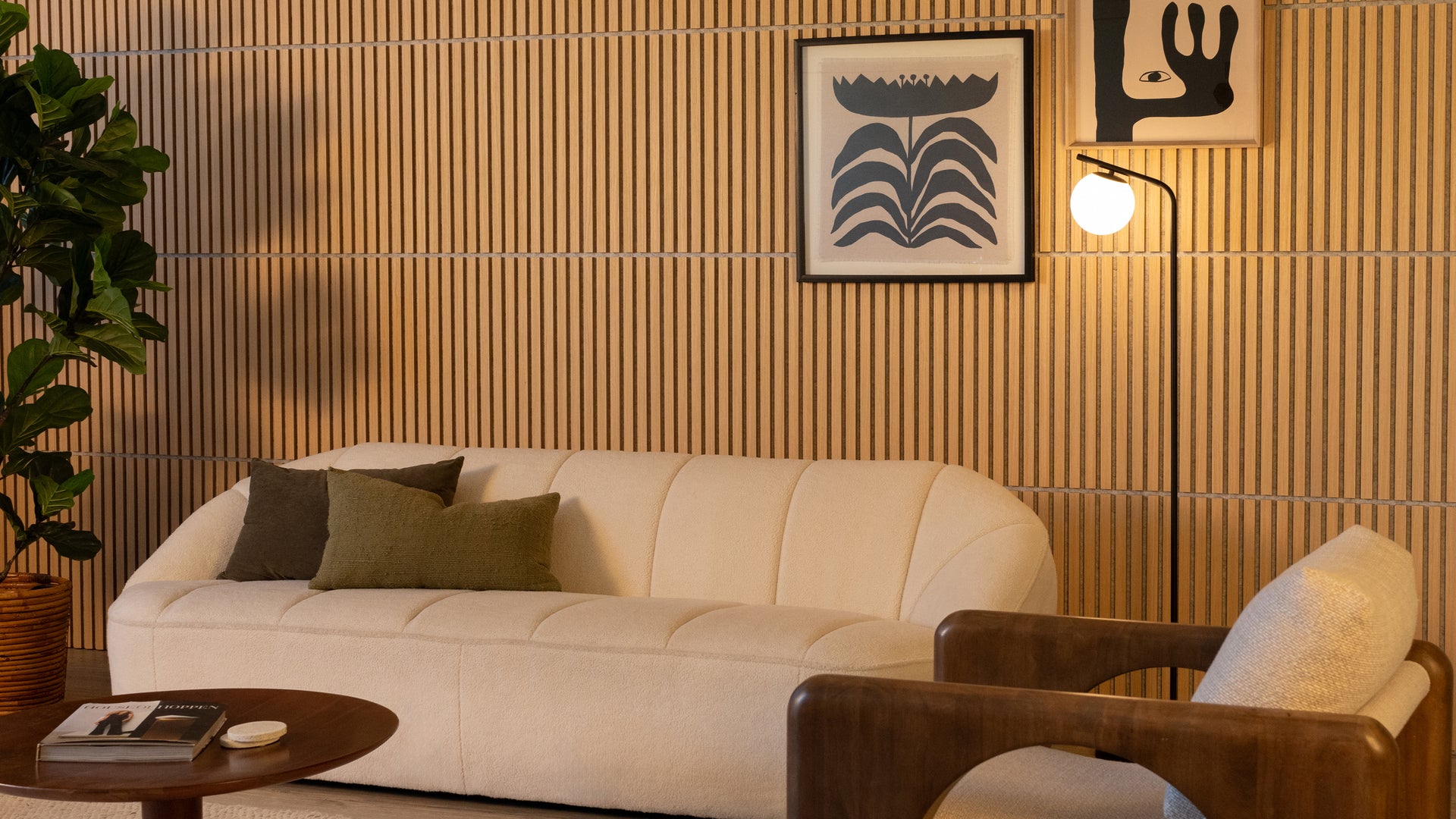 A modern living room with a cream-colored sofa, green and brown pillows, a wooden armchair, a round coffee table, a tall plant, a lamp, and abstract art on a wood-paneled wall.