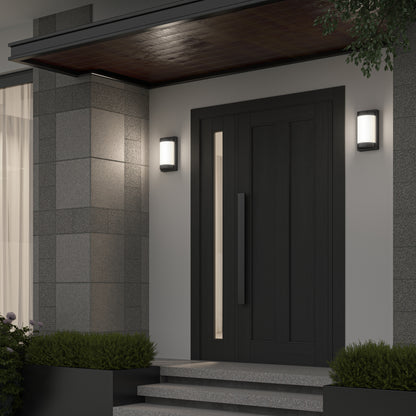 A modern front door with a vertical handle is flanked by two Artika Fogo modern LED indoor-outdoor wall lights, set against gray tiled walls with concrete steps and green shrubs on either side.