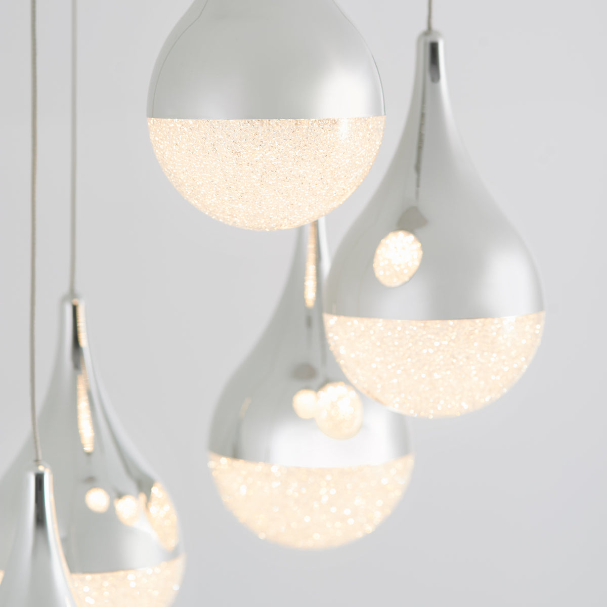 Close-up of modern pendant lights with teardrop shapes, featuring a metallic silver upper half and a textured, sparkling illuminated lower half, against a plain light gray background.