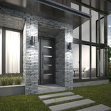 Modern house entry with a dark door featuring horizontal glass panels, surrounded by textured gray stone. Large windows reveal a bright interior with a couch. Concrete stepping stones lead through a grassy lawn.