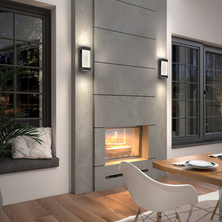 A modern living room features a gray stone fireplace with a cozy fire, two Artika Percy LED indoor-outdoor wall lights, a wooden dining table and chairs, large windows, and a window seat with cushions and a plant.
