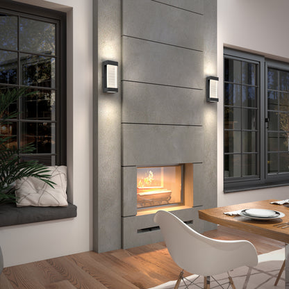 A modern living room features a gray stone fireplace with a cozy fire, two Artika Percy LED indoor-outdoor wall lights, a wooden dining table and chairs, large windows, and a window seat with cushions and a plant.
